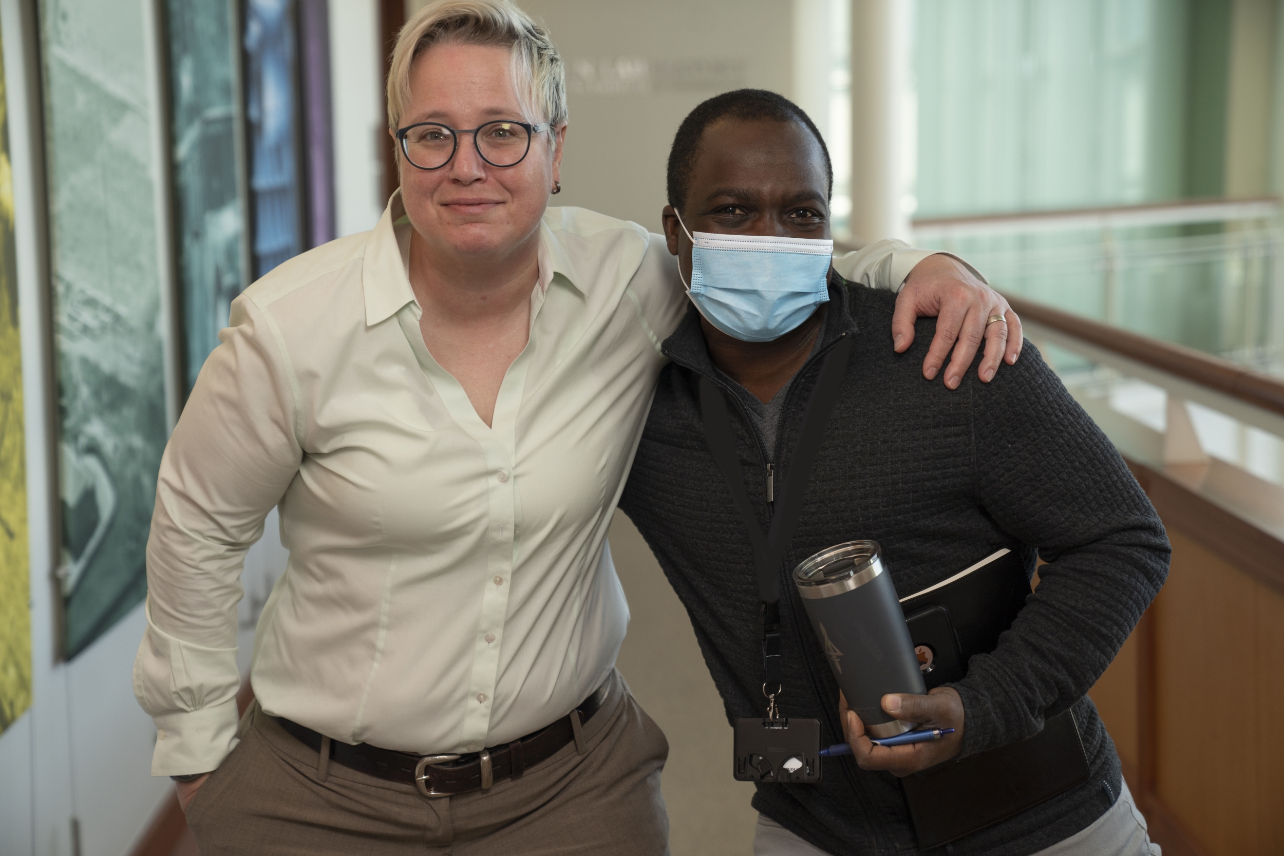 Molly Crane poses smiling for a photo with colleage Raoul Oudraogo in a hallway. To their right is a wall with photos on it, and to their left a balcony that overlooks an atrium.