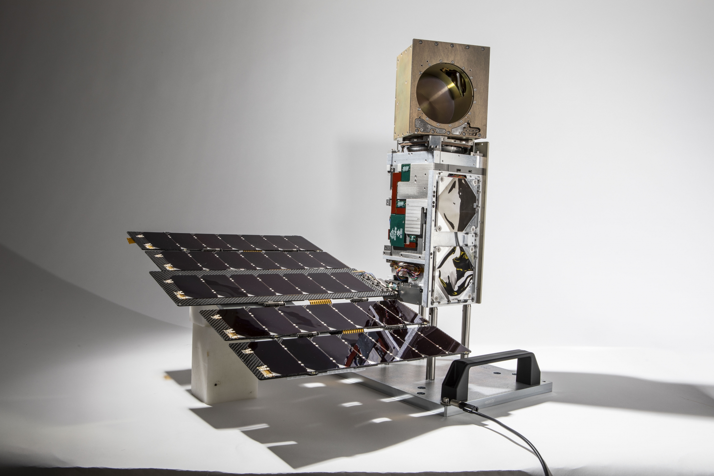 A small satelitte photographed against a white background. About , about 10 cm X 10 cm X 36 cm in size, it consists of a small solar panel array, connected to the satellite bus standing upright, topped with a golden cube. 