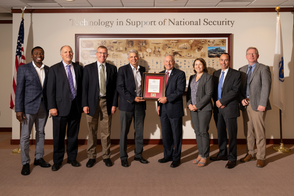 A photograph of eight individuals, with the two in the center holding a plaque in front of a wall that says "Technology in Support of National Security."