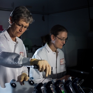 Two women in white lab coats standing with vials in foreground.