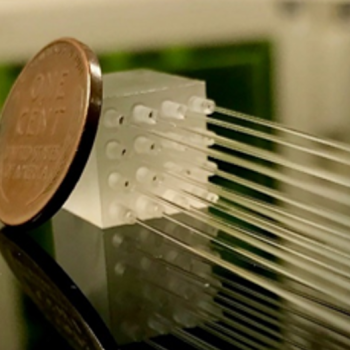 A photo of a small translucent cube with 16 holes in it, from which fiber optics are coming out of. A penny rests against the cube to show its scale.