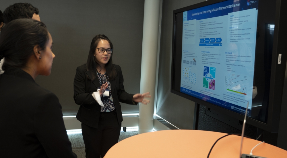 A researcher stands and points to screen showing her research poster. She is talking about the research with two people looking at the screen.