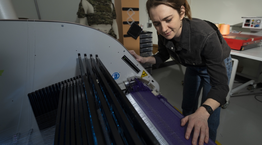 Erin Doran, a textile specialist, is photographed next to a textile weaving machine, with her hands on a fabric being woven. 