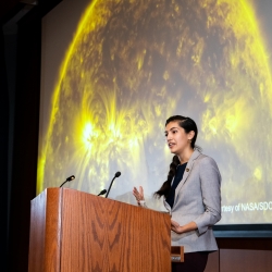 Natalia Guerrero stands at the lecturn and gives her keynote speech in front of a large image of a sun