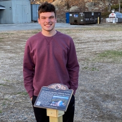 Scout Carter Purple stands outside in front of the plaque he made containing information about the Millstone Hill Radar and Haystack Observatory