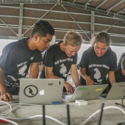 Three students stand together at a table, looking down each at a laptop, discussing. Two other students are in the background. The building they are in looks like an aircraft hanger. 