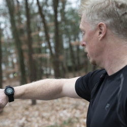 A man in black t-shirt looks at a smartwatch on his wrist, which is displaying a green circle with a thumbs up sign. The man is outside, with trees and leaves in the background.