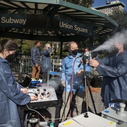 Six scientists in blue lab coats stand in NYC, under a metal gazebo that says "Subway, Union Square" Two of the scientsits operate a small sprayer, on a tripod, that is shooting out a plume of white particles.