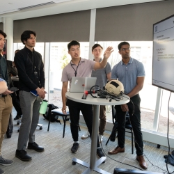 A group of cadets and staff look at a large monitor as a cadet presents his AI casualty status tool.